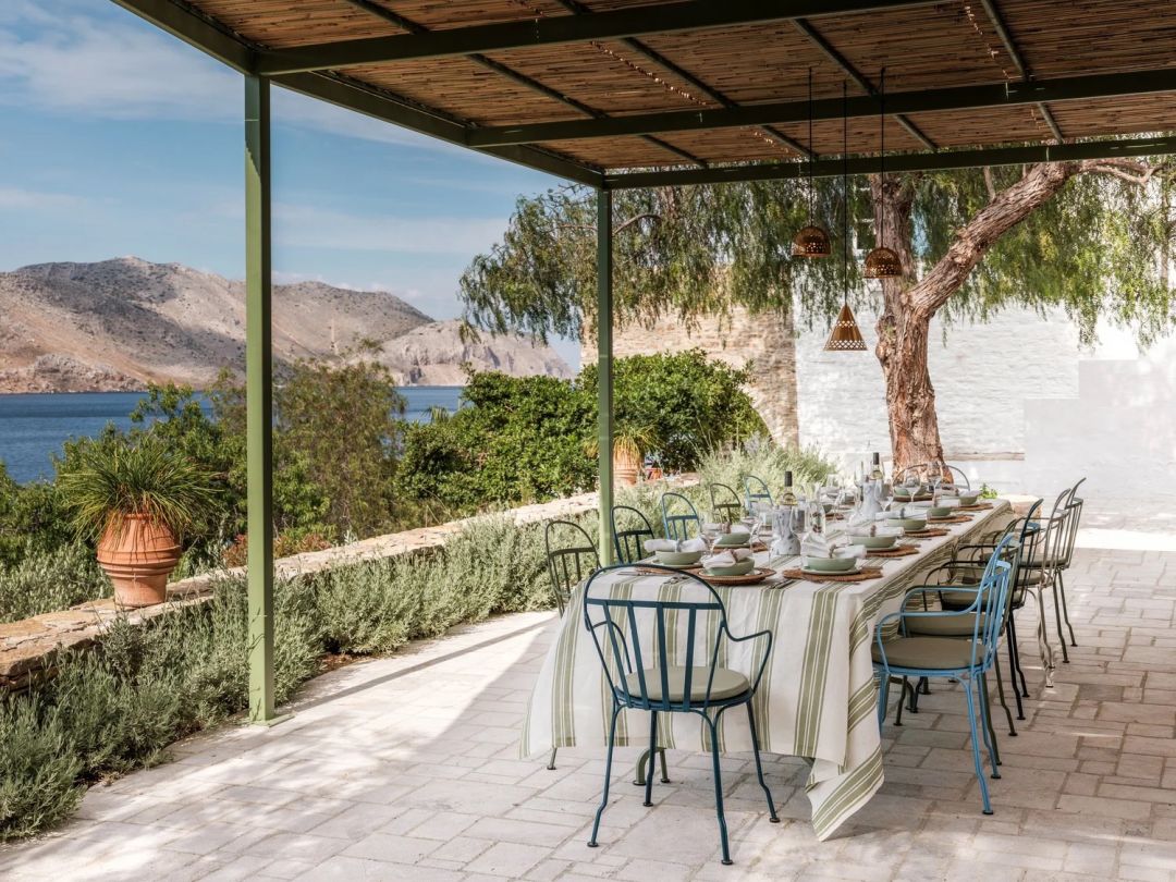 A long lunch table under a reed-roofed pergola at The Selene, Symi, laid with striped linen and wirework chairs, the bay beyond.
