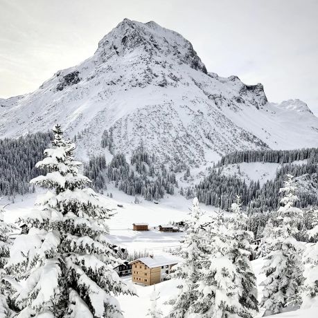 The Sonnenhof in its winter landscape above Lech, a cluster of dark timber houses in deep snow beneath the Omeshorn, snow-laden spruces framing the valley.