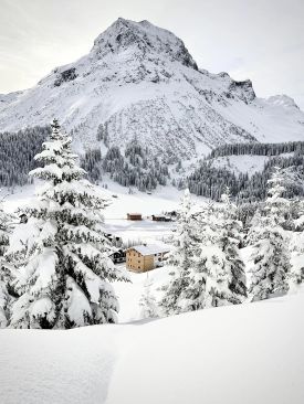 The Sonnenhof in its winter landscape above Lech, a cluster of dark timber houses in deep snow beneath the Omeshorn, snow-laden spruces framing the valley.