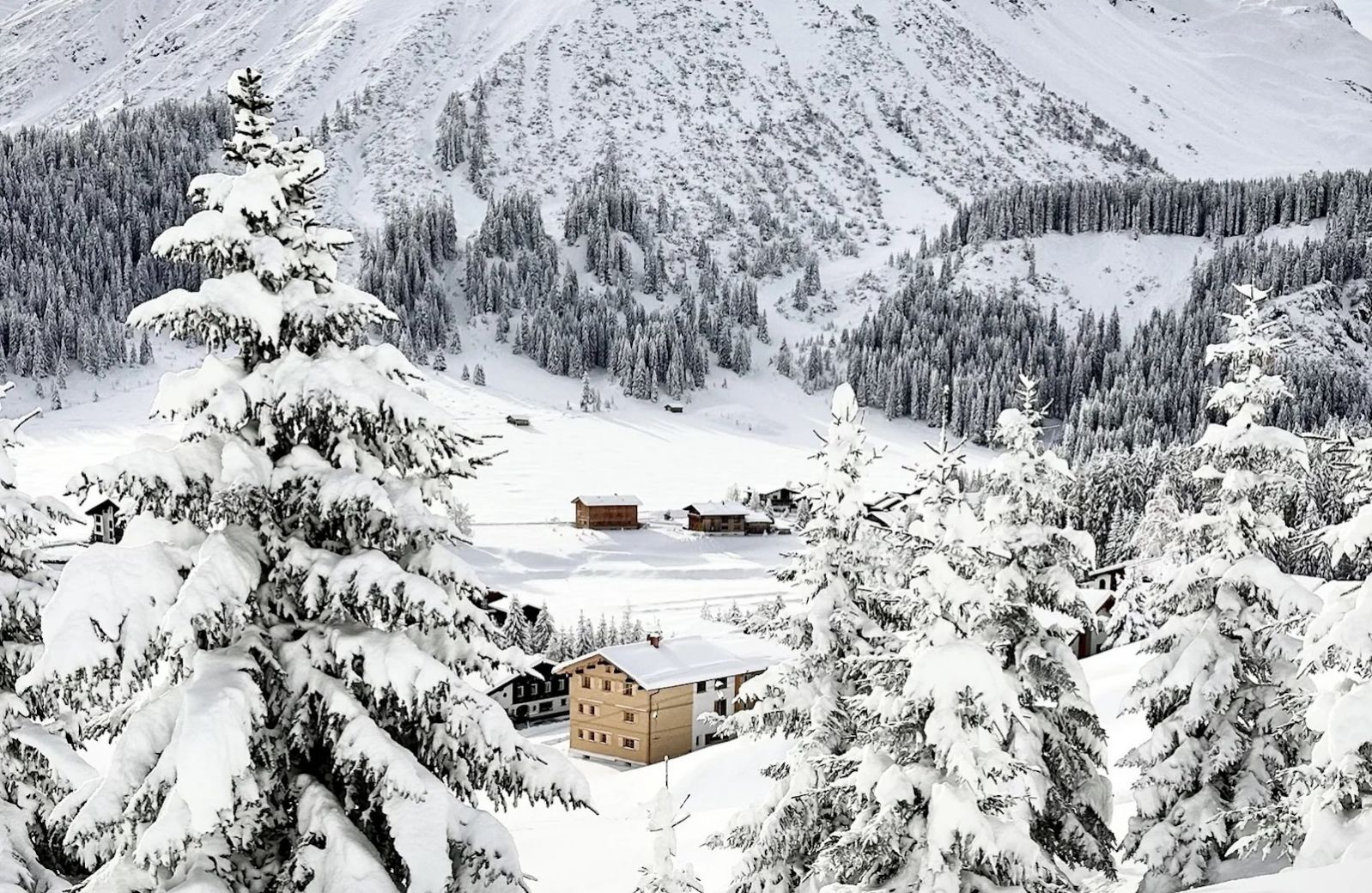 The Sonnenhof in its winter landscape above Lech, a cluster of dark timber houses in deep snow beneath the Omeshorn, snow-laden spruces framing the valley.