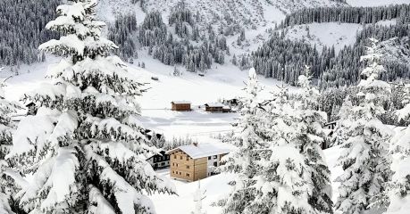 The Sonnenhof in its winter landscape above Lech, a cluster of dark timber houses in deep snow beneath the Omeshorn, snow-laden spruces framing the valley.