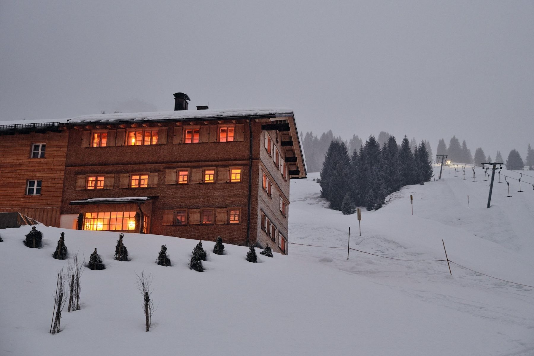The Sonnenhof at dusk in falling snow above Lech, warm light in every window across three floors of the Walser house, the Hinterwies drag lift climbing the slope alongside.