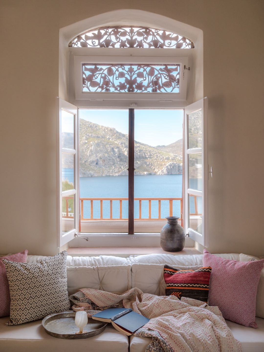 An arched window with carved fretwork fanlights opens onto Nimborio Bay at The Selene, Symi, a cushioned window seat below.|Photo ©️ Will Slater