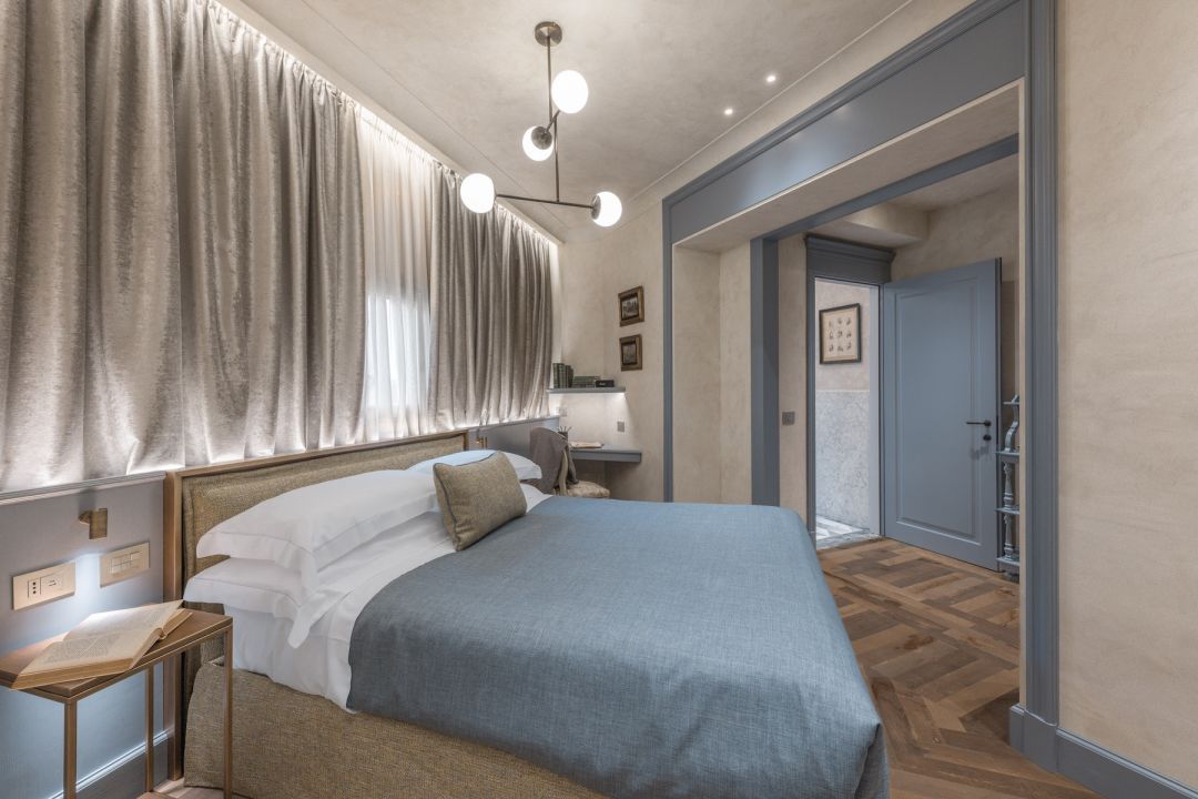 A suite at Santa Croce Historical Home with a pale blue bedcover, a headboard wall hung in lit silver-grey drapery, blue-grey door frames and herringbone parquet leading to a marble bathroom.
