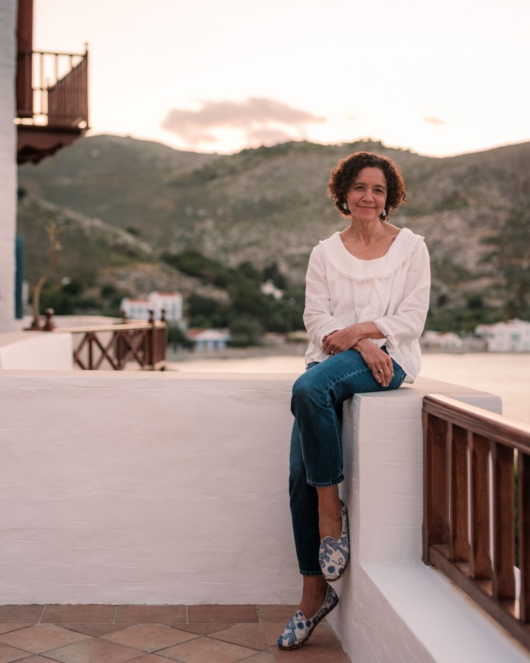 Annette Thomas, owner of The Selene, seated on a whitewashed terrace wall above Nimborio Bay, Symi.|Photo ©️ Will Slater