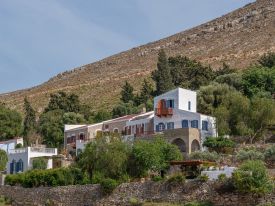 The Selene on Nimborio Bay, Symi: a whitewashed tower with blue shutters beside a stone wing, above a dry-stone sea wall and bare mountainside.| Photo ©️ Will Slater