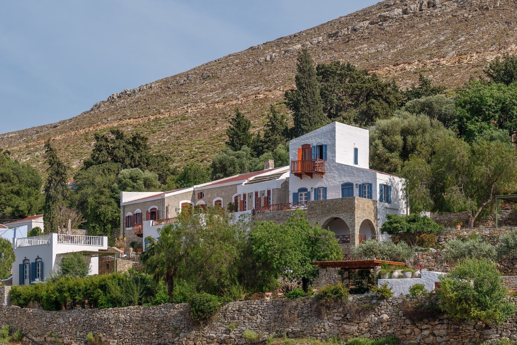 The Selene on Nimborio Bay, Symi: a whitewashed tower with blue shutters beside a stone wing, above a dry-stone sea wall and bare mountainside.|Photo ©️ Will Slater