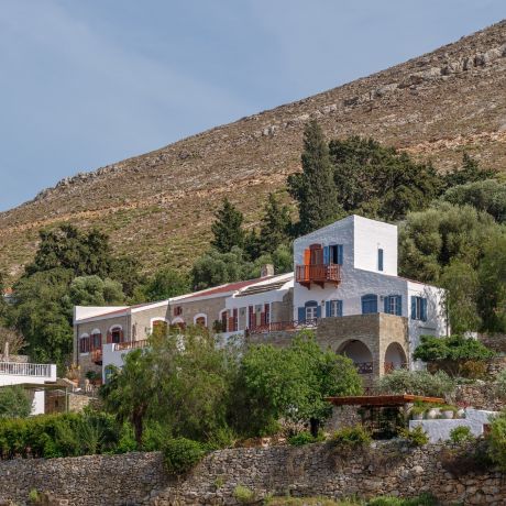 The Selene on Nimborio Bay, Symi: a whitewashed tower with blue shutters beside a stone wing, above a dry-stone sea wall and bare mountainside.| Photo ©️ Will Slater