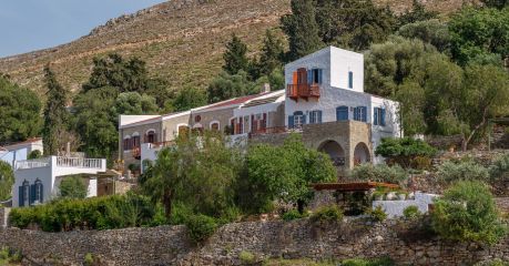 The Selene on Nimborio Bay, Symi: a whitewashed tower with blue shutters beside a stone wing, above a dry-stone sea wall and bare mountainside.| Photo ©️ Will Slater