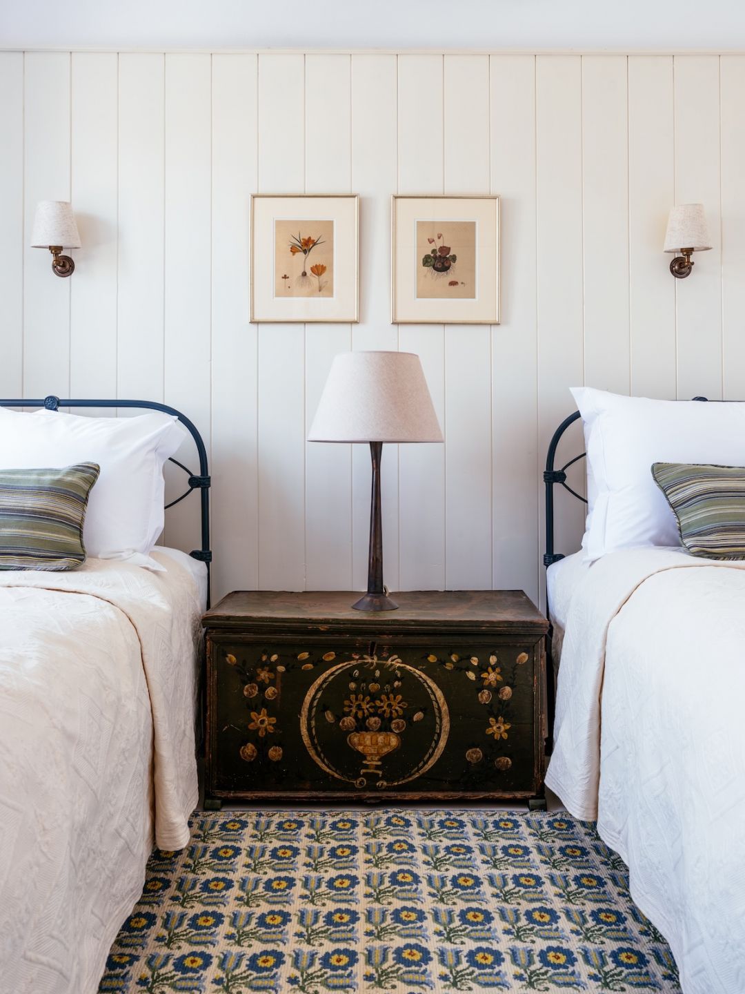 Twin iron beds either side of a painted dowry chest at The Selene, Symi, with brass sconces, botanical prints and a floral rug.|Photo ©️ Will Slater