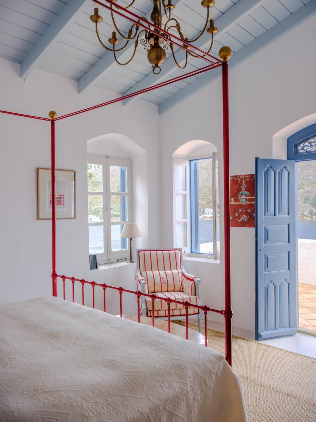 A red-painted iron four-poster under a pale blue beamed ceiling at The Selene, Symi, blue panelled doors open to the terrace.