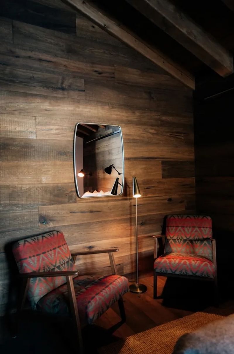 A corner of L'Ovella Negra with a reclaimed oak plank wall, rounded mirror, brass floor lamp and two mid-century armchairs in red patterned wool. Caption: Reclaimed oak, brass and a flash of pattern.