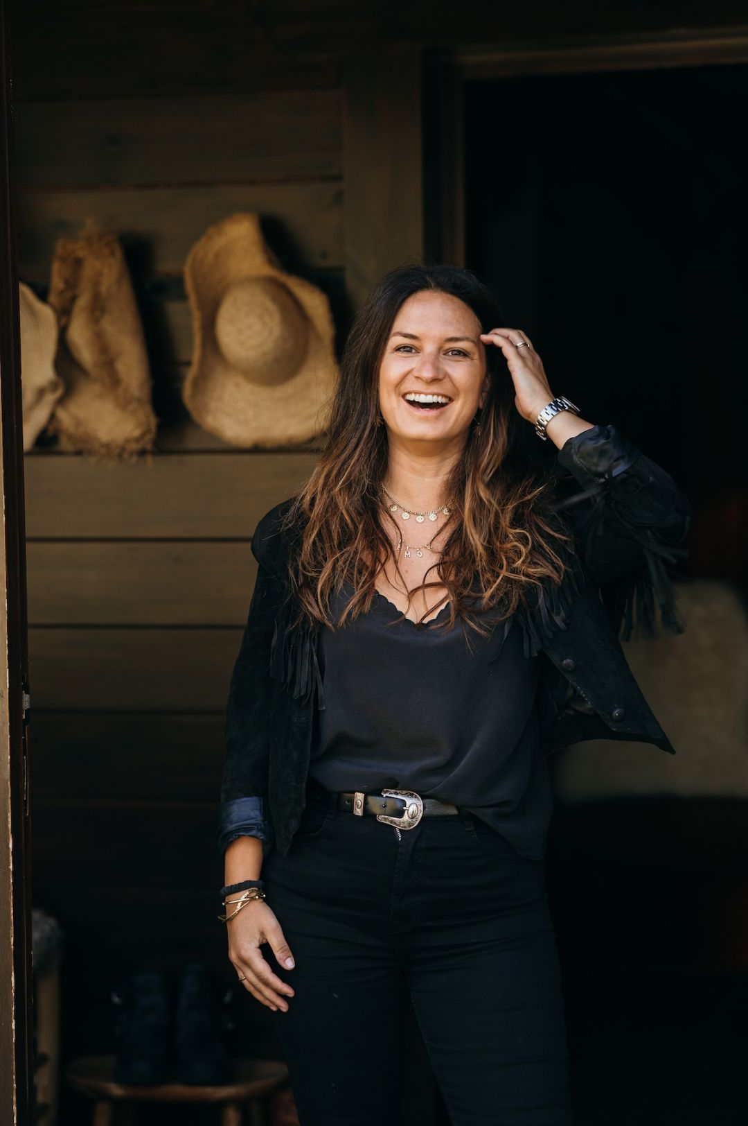 Prisca Llagostera, owner of L'Ovella Negra, laughing in a black fringed jacket in the doorway of the lodge, straw hats hanging behind her. Caption: Prisca Llagostera, the black sheep herself.