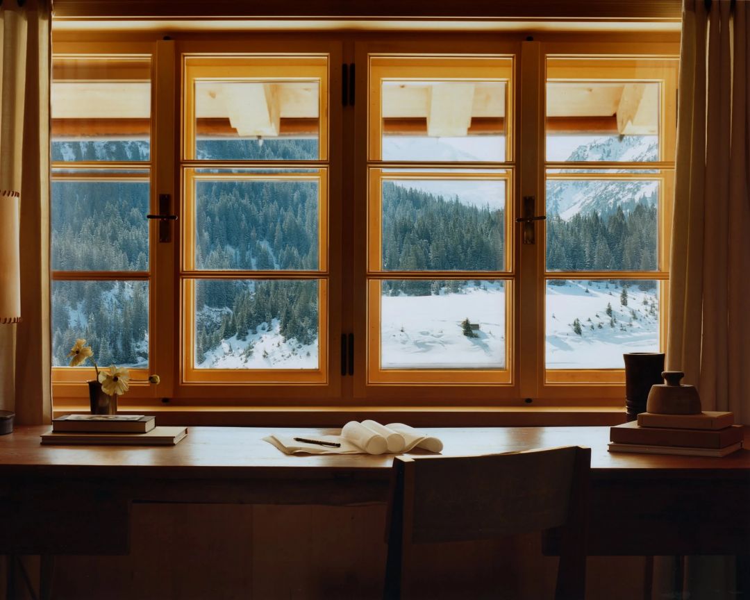 A writing desk at the Sonnenhof set beneath a box casement window in pale spruce, an open book and a small vase of flowers, snow-covered forest and slope beyond the glass.