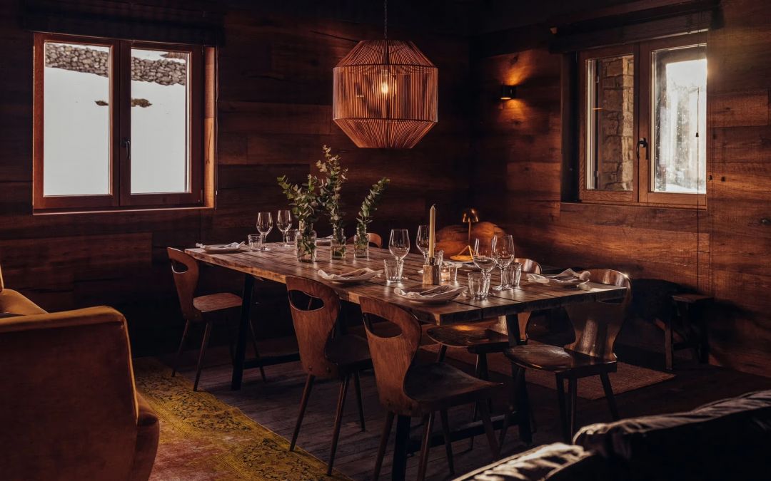 The Lodge Canteen at L'Ovella Negra laid for dinner, timber table, moulded plywood chairs, a woven pendant above and snow banked against the windows. Caption: Dinner in the Canteen, snow to the windowsills.