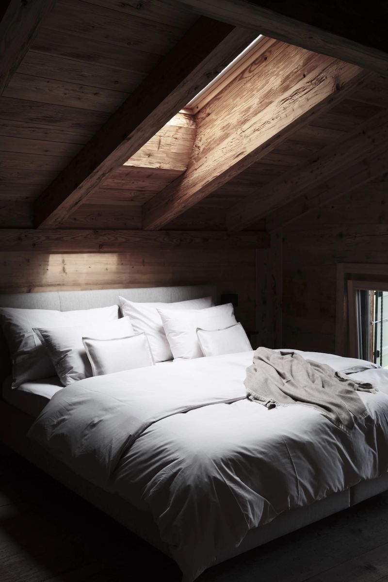 An attic bedroom at Haus Mannenberg tucked beneath the sloping timber roofline, a recessed skylight drawing daylight down the raw wood, the bed dressed in crisp white linen with a soft knitted throw folded across it.