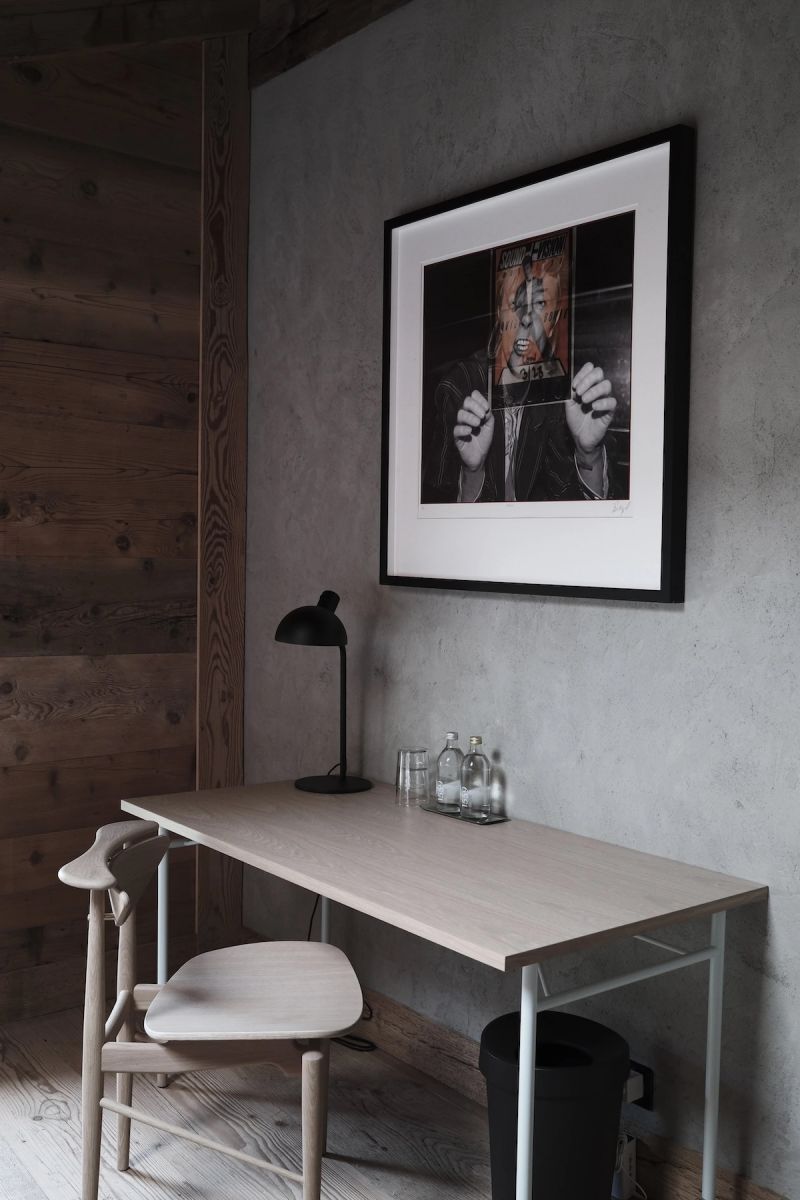 A quiet work corner in a Haus Mannenberg guest room: a pale oak desk and light wood chair against a timber-plank and grey-plaster wall, a black task lamp and glass water bottles on the desk, and a black-framed portrait photograph above.