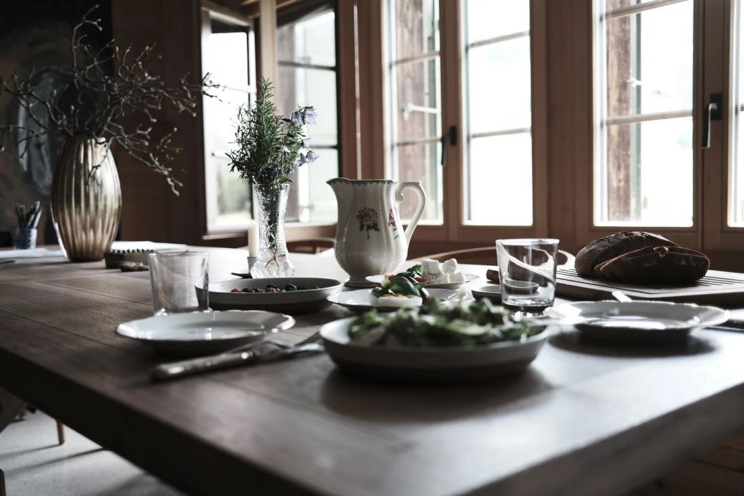 The communal breakfast table at Haus Mannenberg by a run of timber-framed windows, laid with white plates, a floral jug, cut garden flowers and a dark country loaf, morning light falling across the dark wood.