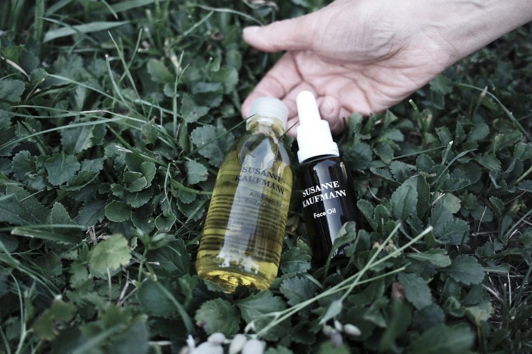 A hand reaching for two Susanne Kaufmann oils, an amber-glass face oil and a clear arnica body oil, resting among green grass and clover in the grounds at Haus Mannenberg.