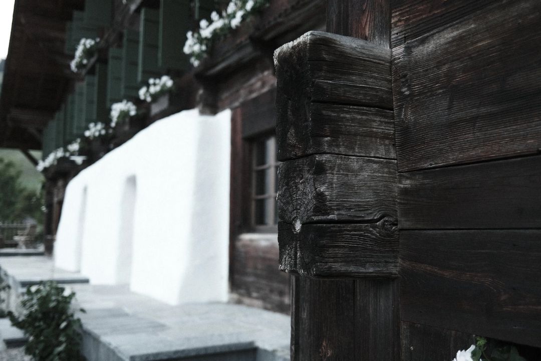A close detail of Haus Mannenberg's weathered, near-black timber corner beams, the deep grain and joinery in sharp focus, while a whitewashed wall, green shutters and white geraniums recede softly behind.