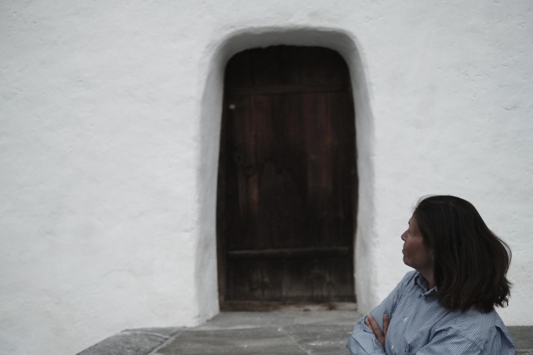 Maria, co-owner of Haus Mannenberg, sitting outside against a whitewashed wall beside a deep-set antique timber door, arms folded, in a blue striped shirt, looking off to one side.
