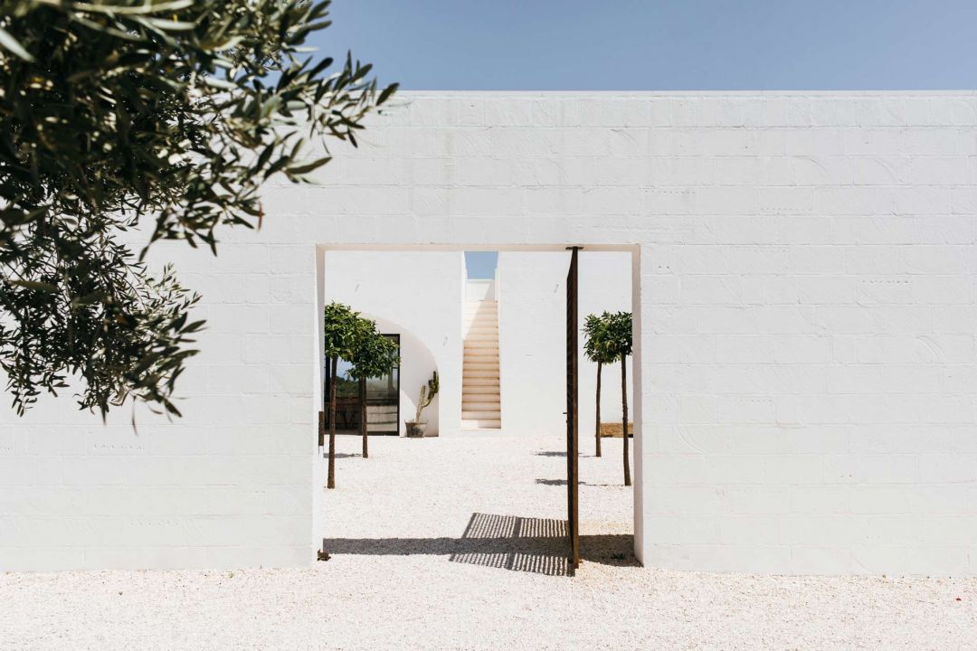 The single pivoting gate set into the whitewashed perimeter wall of Masseria Moroseta, opening onto the gravel courtyard, potted citrus and pale staircase, Ostuni, Puglia | The Aficionados.