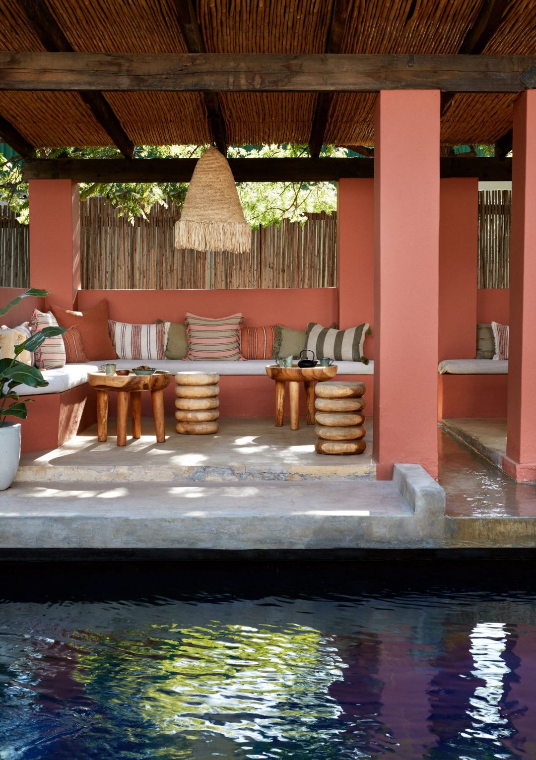 The terracotta cabana at Kokos Huis seen across the pool, a fringed raffia pendant hanging beneath the reed roof above plaster banquettes in striped cushions and carved timber stools.