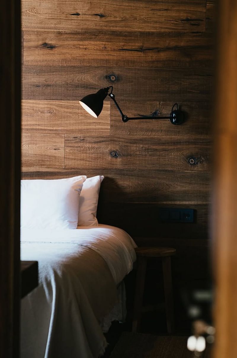 A bedroom at L'Ovella Negra, a black articulated wall lamp on a dark oak plank wall above crisp white linen and a wooden stool. Caption: Dark oak, white linen, and light exactly where you want it.