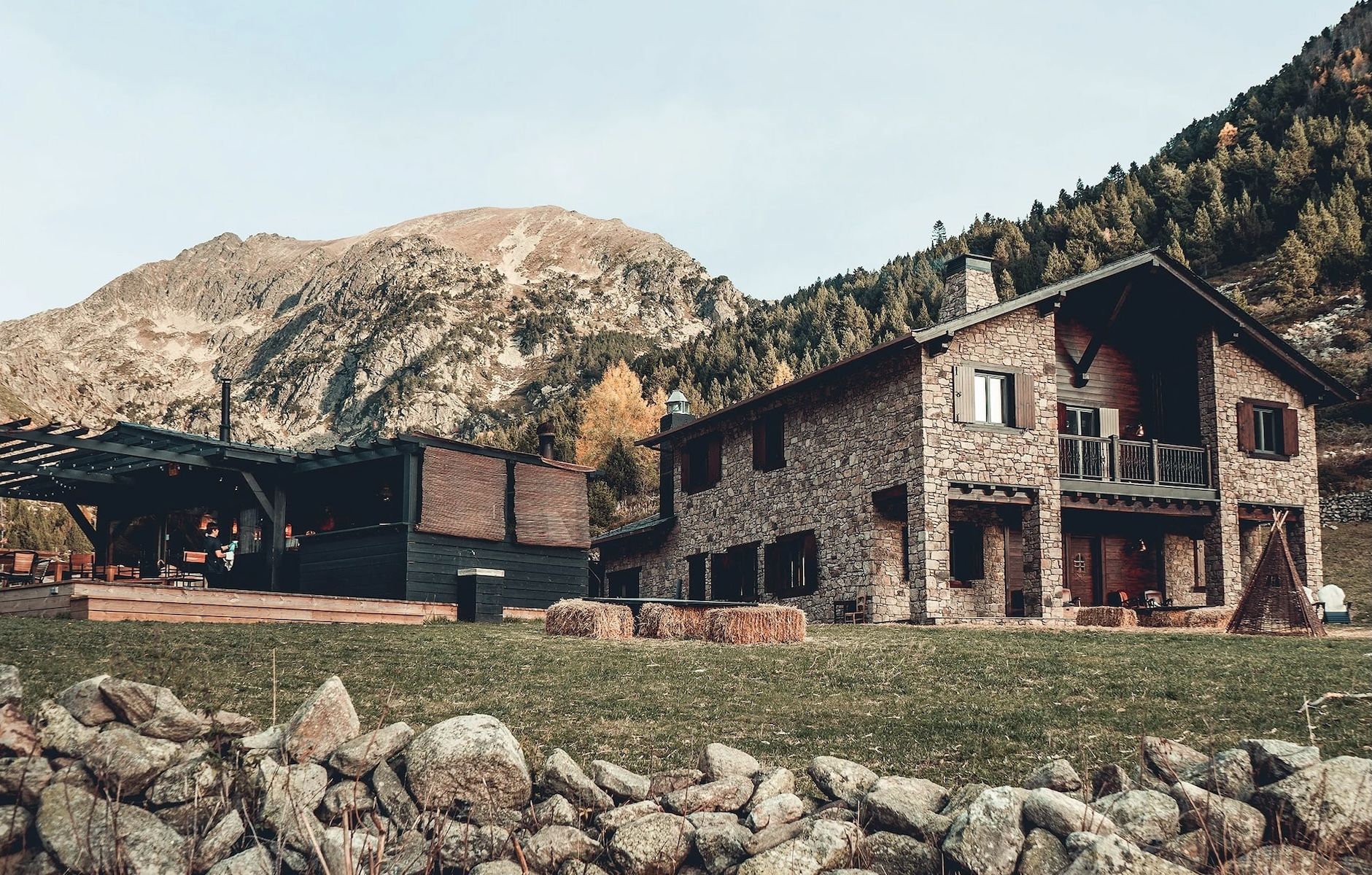 The stone and timber exterior of L'Ovella Negra in the Vall d'Incles, Andorra, with the open-sided pavilion, festoon lights and hay bales on the grass. Caption: The lodge and its pavilion at the head of the valley, hay bales still on the grass.