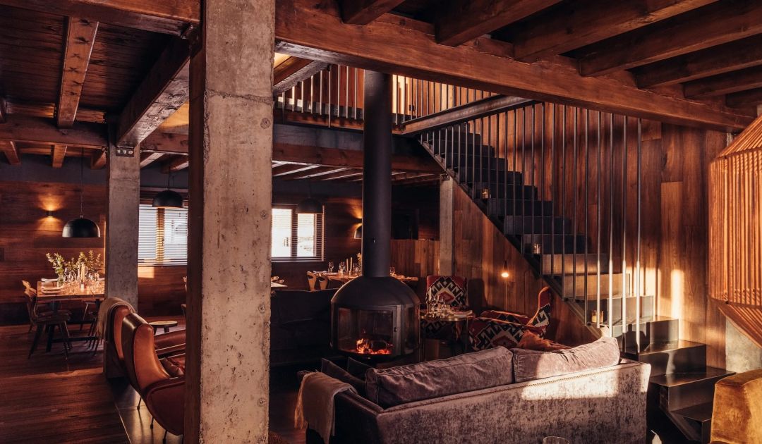 The main room at L'Ovella Negra, a suspended black stove and flue between concrete pillars and old timber beams, velvet sofa, leather chairs and a steel stair. Caption: The suspended chimney, the object that holds the whole idea of the place.