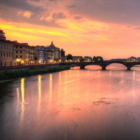 The Arno at sunset from the Lungarno, an arched stone bridge spanning the river between lines of riverside palazzi, their lights reflected in the water beneath a pink and gold sky.