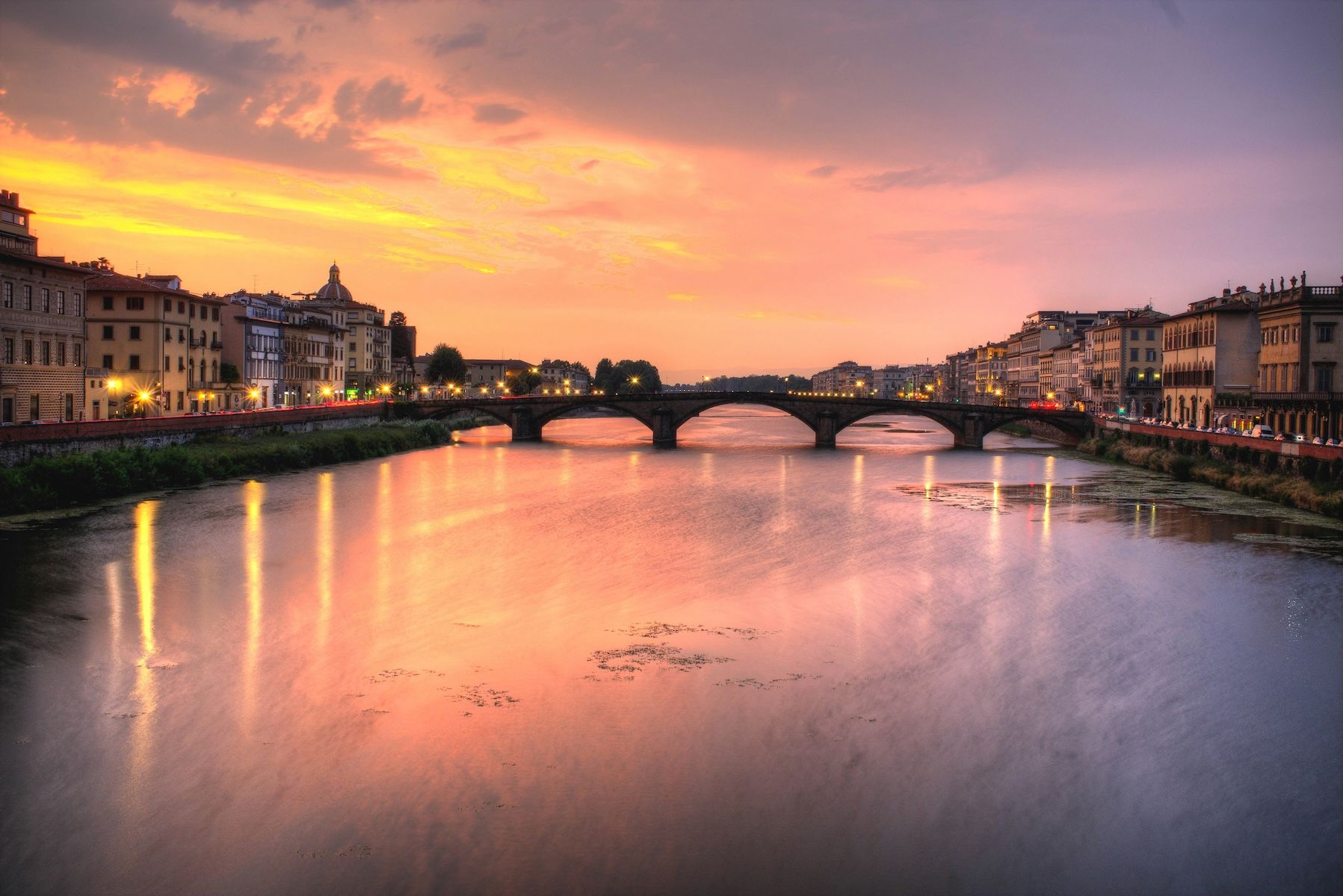 The Arno at sunset from the Lungarno, an arched stone bridge spanning the river between lines of riverside palazzi, their lights reflected in the water beneath a pink and gold sky.