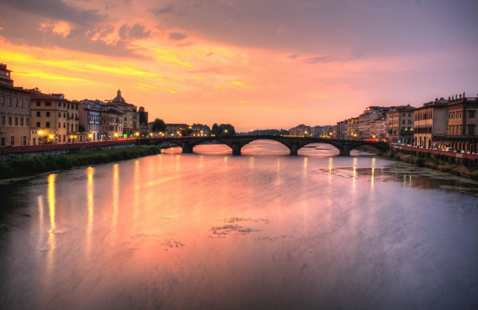 The Arno at sunset from the Lungarno, an arched stone bridge spanning the river between lines of riverside palazzi, their lights reflected in the water beneath a pink and gold sky.