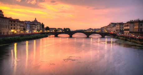 The Arno at sunset from the Lungarno, an arched stone bridge spanning the river between lines of riverside palazzi, their lights reflected in the water beneath a pink and gold sky.