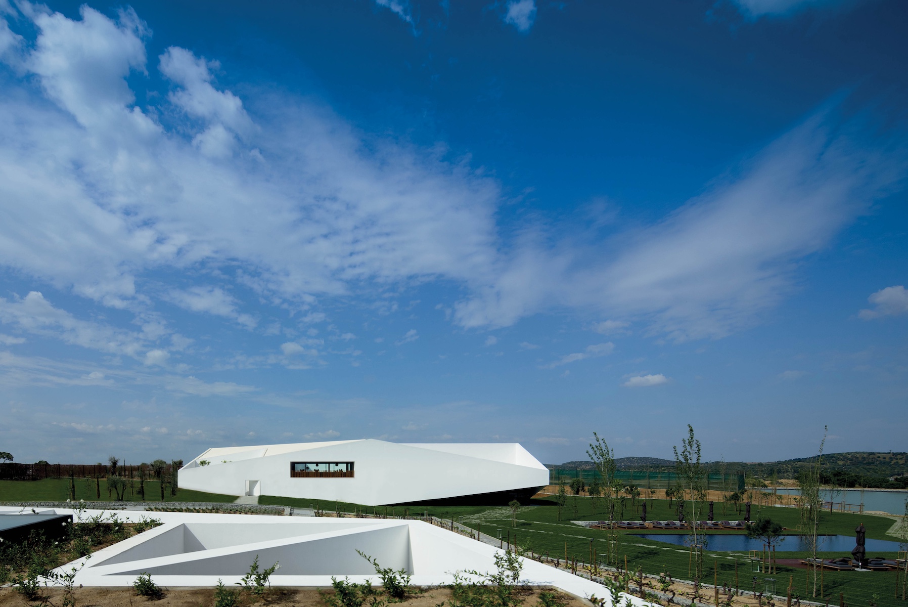 The sculptural white prism of L'AND Vineyards rising above lawns and a reflecting pool in the Alentejo, Portugal, its sheared roofline set against a wide blue sky.