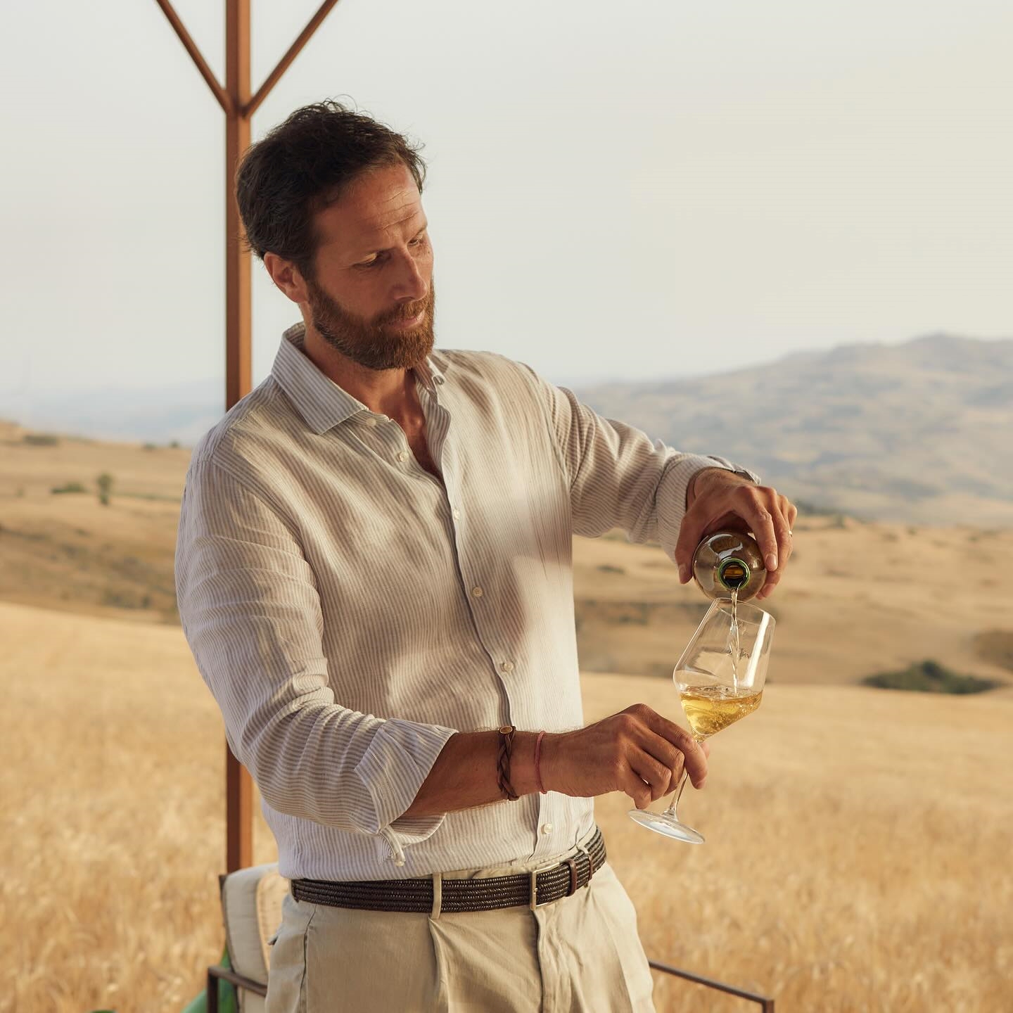 Manfredi Rizzuto | Amna in white shirt pouring a glass of local wine outdoors agains t agolden field of crops on the island of Sicily Italy | Sasufa Hotel | Manmade: 12 Hotels Shaped by Men of Design and Style