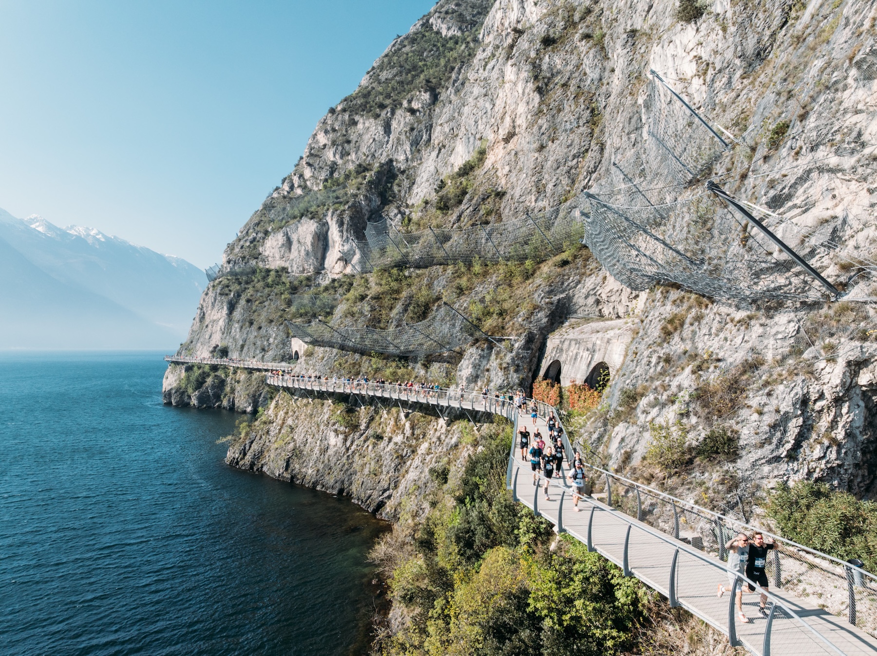 Runners strung along the cantilevered cliff cycleway at Limone sul Garda during PEAK Lake Garda 42, blue water on one side and sheer rock on the other.