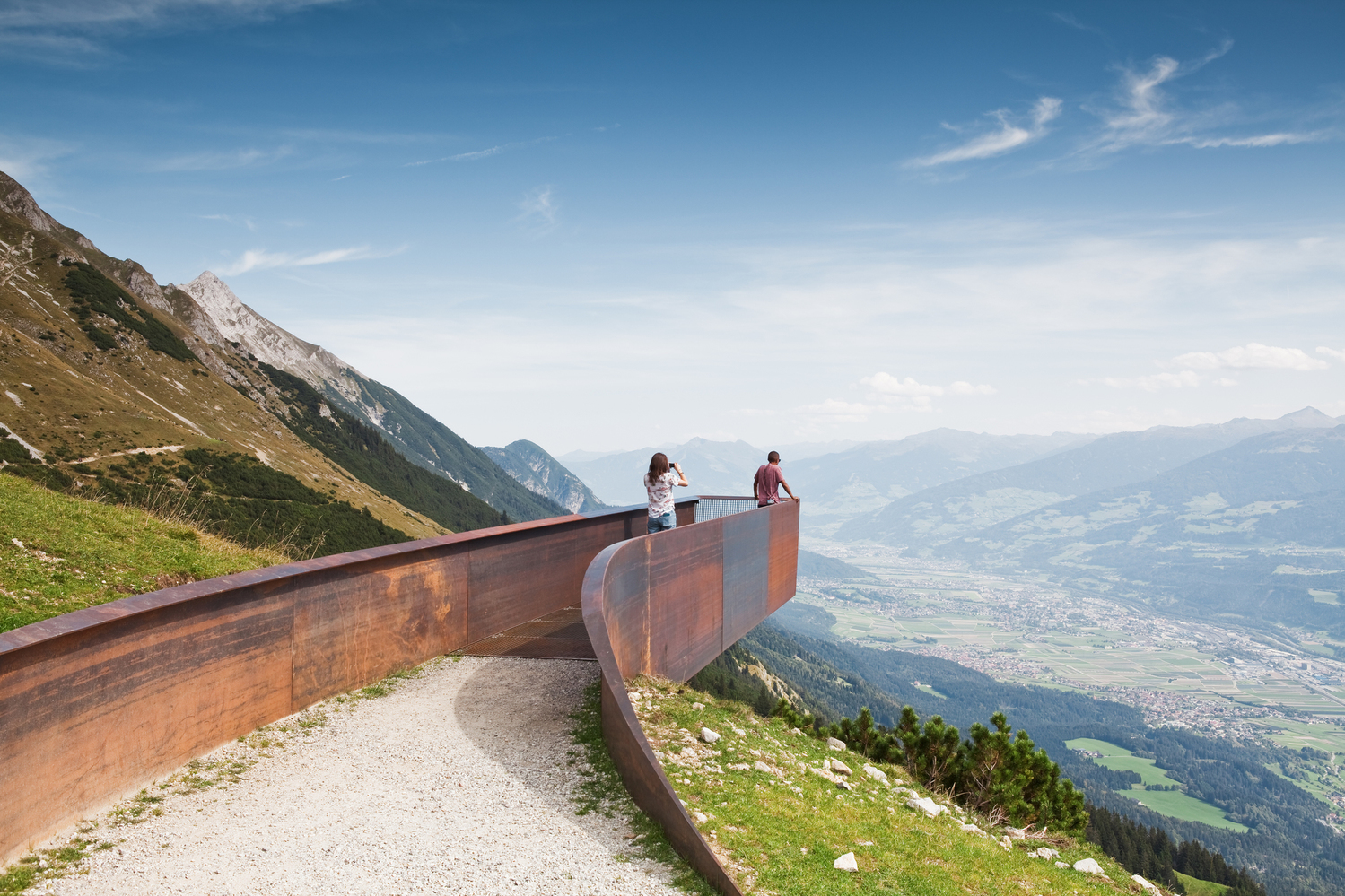 Two visitors at the end of Snøhetta’s weathering-steel lookout on the Path of Perspectives, the Nordkette ridge above Innsbruck curving out over the Inn valley | The Aficionados