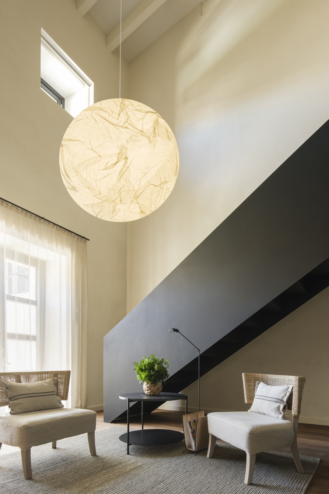 A double-height living space at Can Ferrereta, Mallorca, with a paper-moon pendant, a black steel staircase cutting the wall and cane armchairs below.