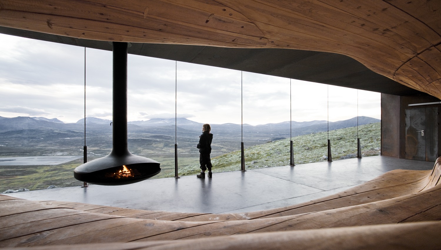A visitor at the glass wall of Snøhetta’s Wild Reindeer Pavilion at Hjerkinn, carved pine curving around a hanging fireplace and the Dovrefjell plateau beyond | The Aficionados