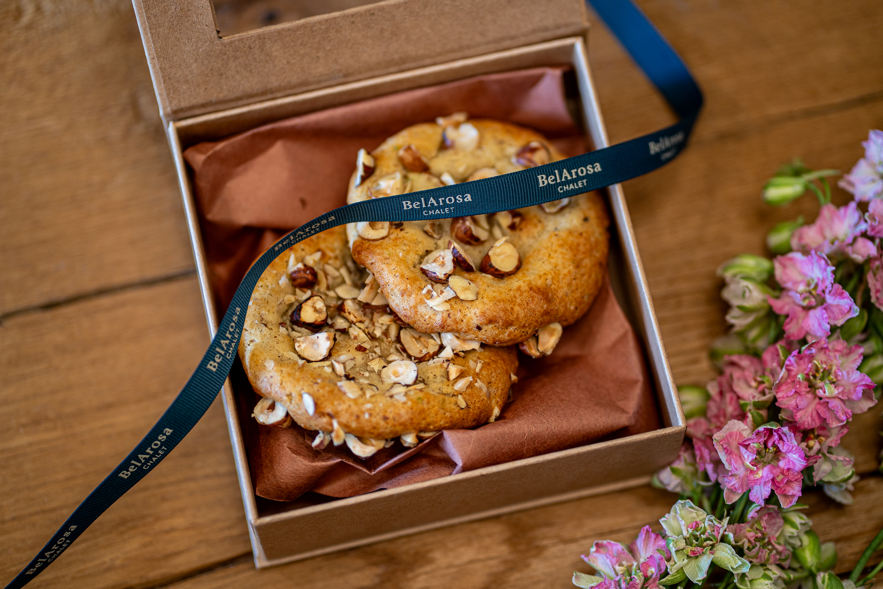 Two BelArosa Chalet honey cookies topped with hazelnuts, in a kraft gift box tied with a navy BelArosa Chalet ribbon, beside pink flowers on wood.