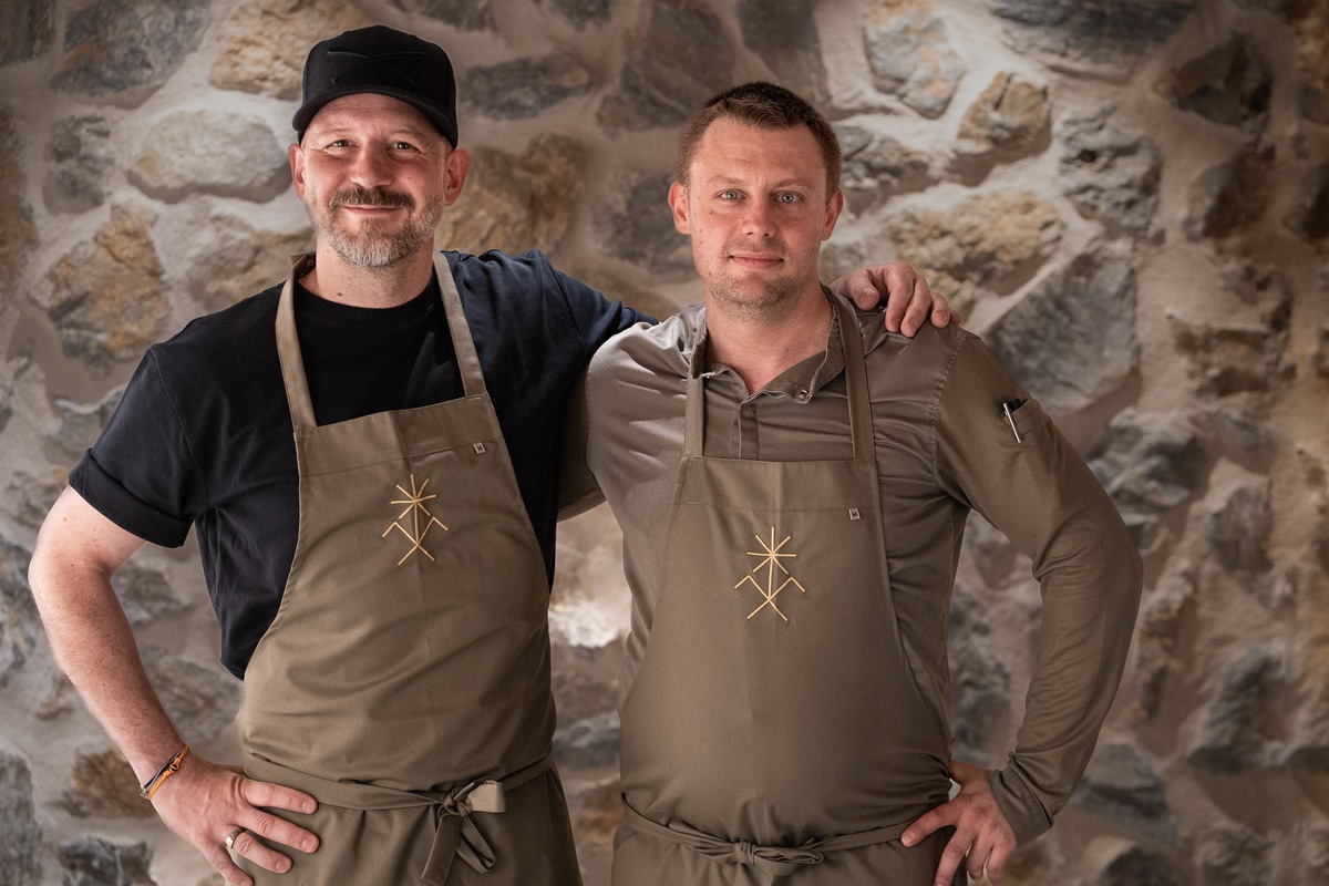 Patissier Christian Hümbs with a fellow chef at the BelArosa Chalet, both in aprons bearing the chalet's Walser house mark, against a stone wall.
