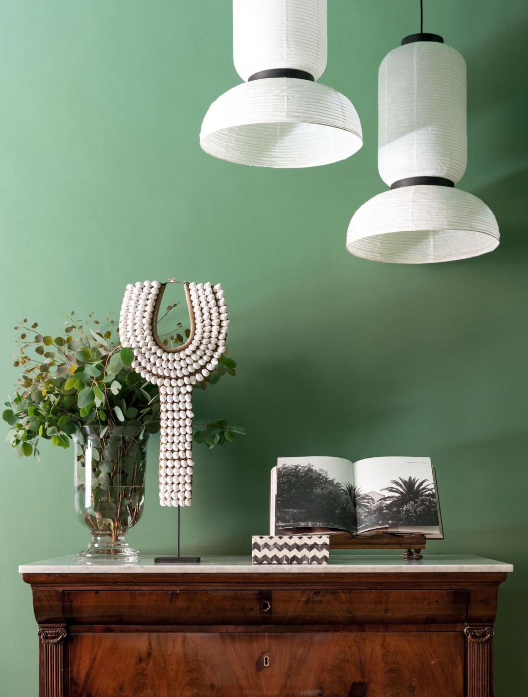 Still-life detail of white paper lanterns, a shell necklace and an antique walnut chest against a sage green wall