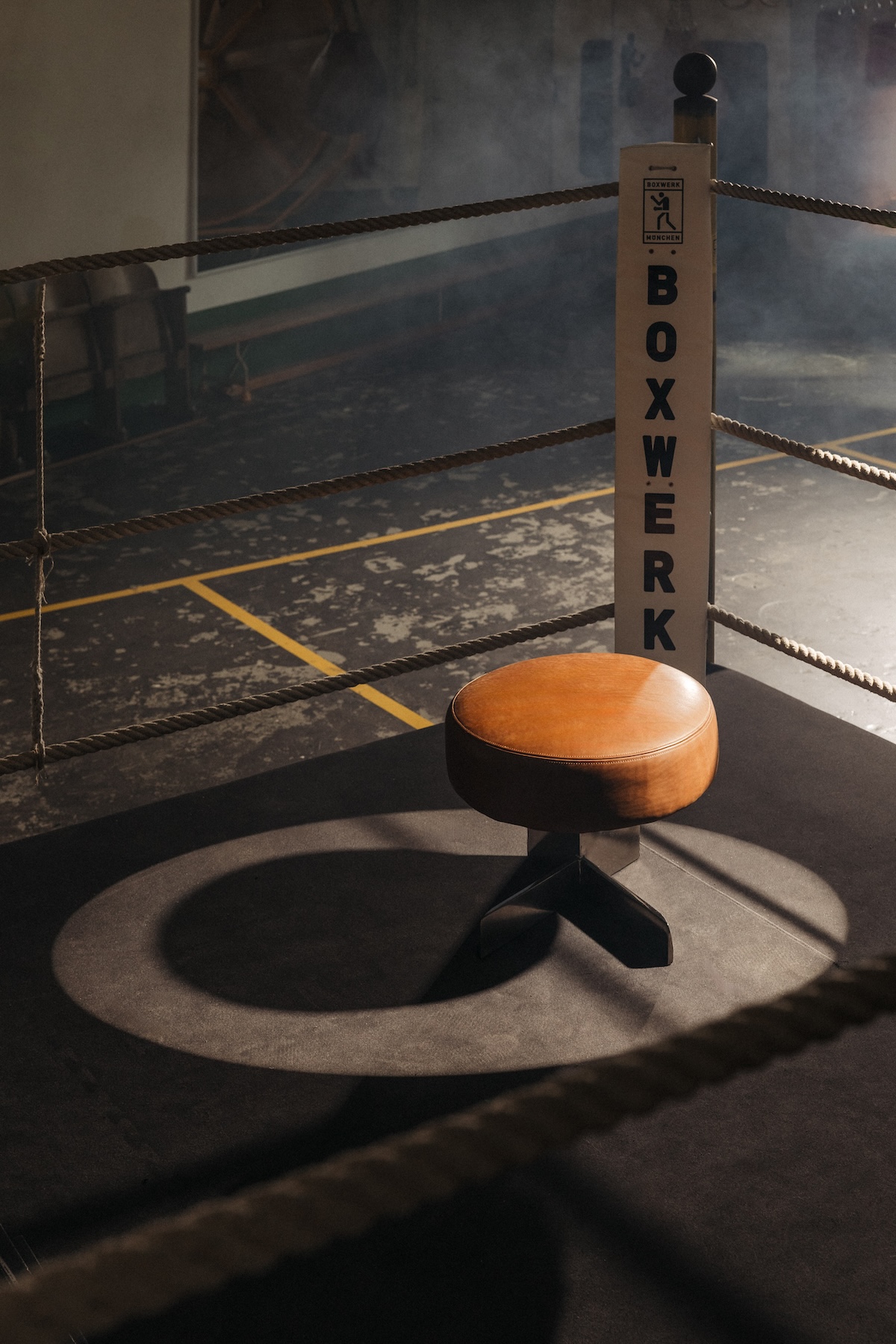 A tan leather Egetemeier stool with a black cross base lit in the corner of a Boxwerk München boxing ring, ropes and haze around it.