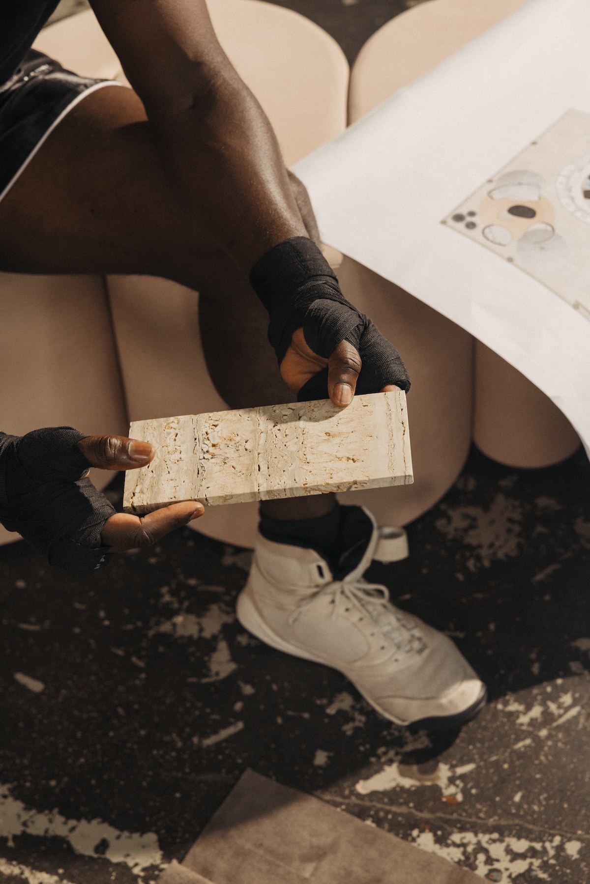 Close-up of a boxer with black hand wraps seated on a curved cream lounge, holding a rectangular travertine stone sample above a paint-flecked studio floor.