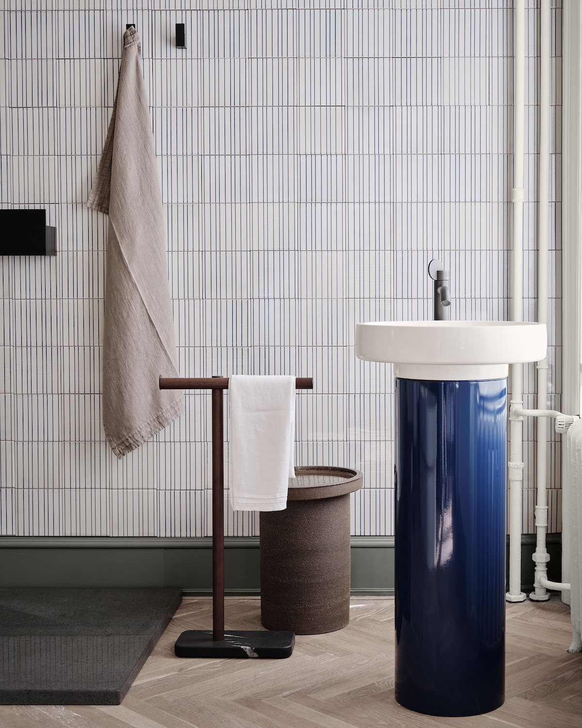 Agape bathroom detail: round white basin on a deep blue gloss pedestal beside a walnut and marble towel stand, set against vertical blue-striped File Under Pop tiles.