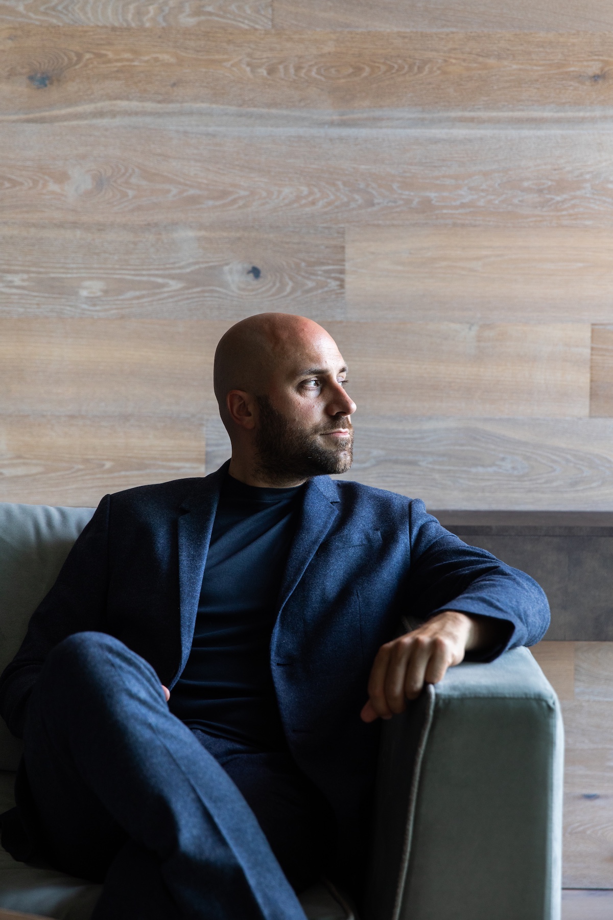 Alessandro Sironi | elegant man sitting wearing a blue suit, relaxed against a grey blue sofa and marble tiled wall | Owner of Filario Hotel Lake Como | Manmade: 12 Hotels Shaped by Men of Design and Style