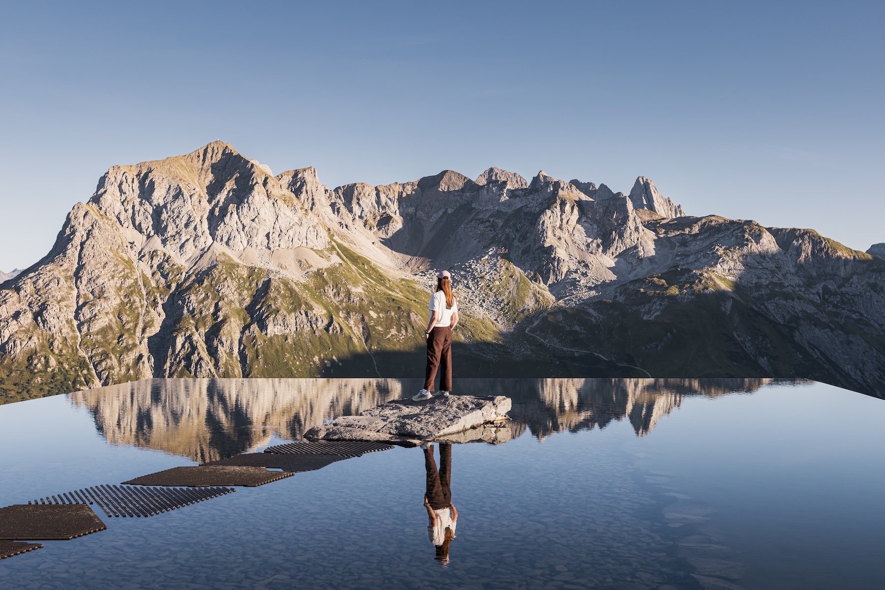 A woman stands on the rock at the centre of Snøhetta’s Lech Mountain Mirror, the Arlberg peaks reflected upside down in the still rooftop pool above Zug | The Aficionados