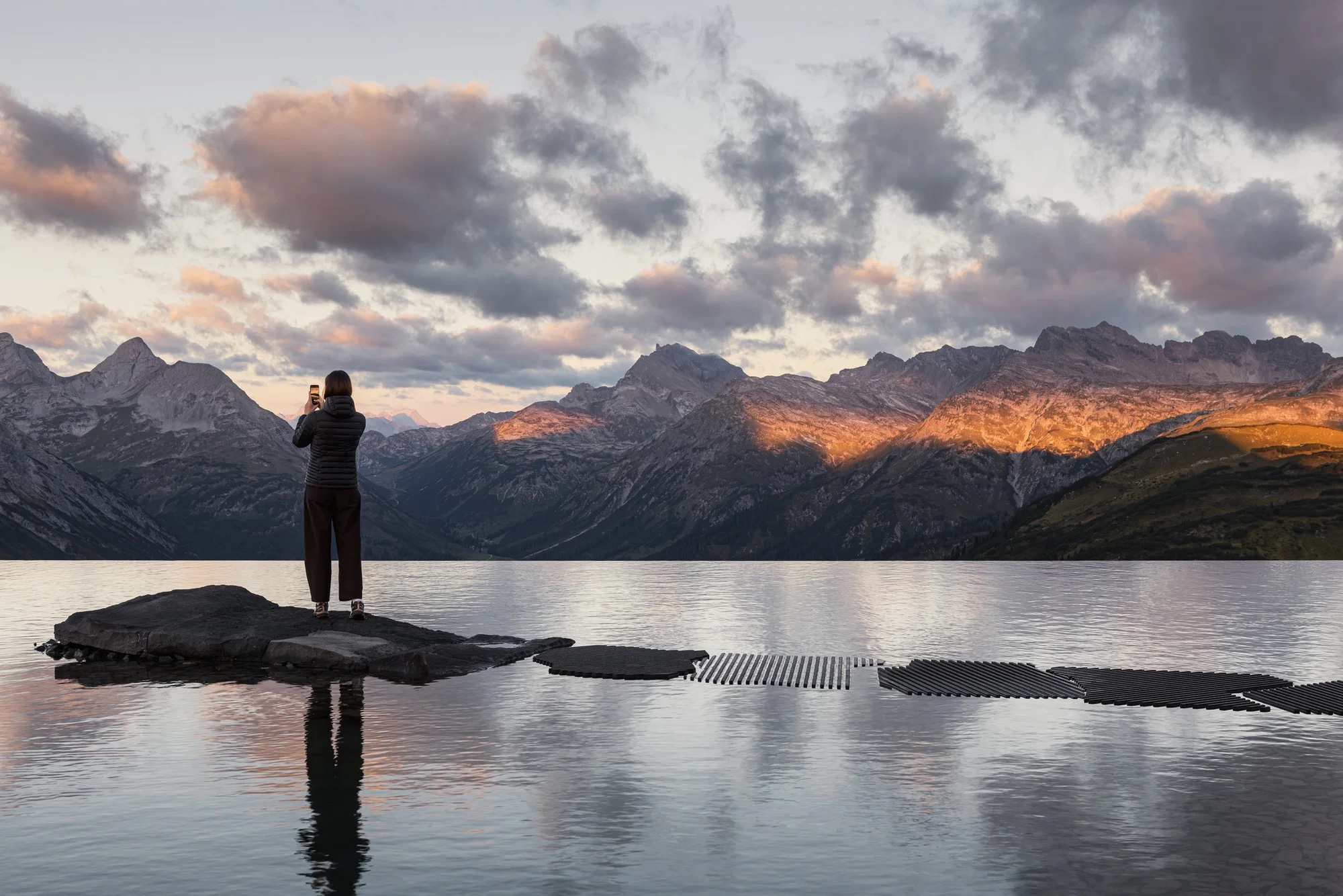 Evening: Snøhetta’s Mountain Mirror A visitor photographs the sunset from the rock in Snøhetta’s Lech Mountain Mirror, stepping stones crossing the water and the last light on the Arlberg peaks | The Aficionados