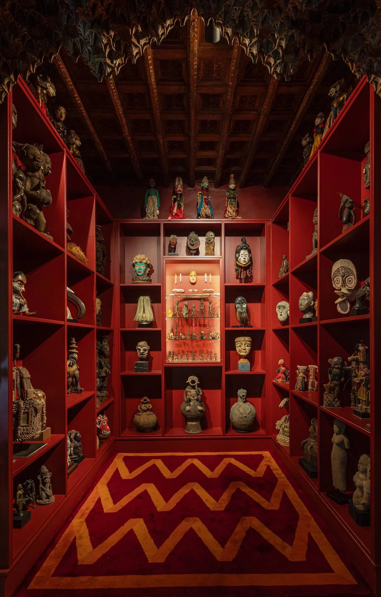 A red-lined display room of carved figures and masks beneath a painted timber ceiling | The Aficionados